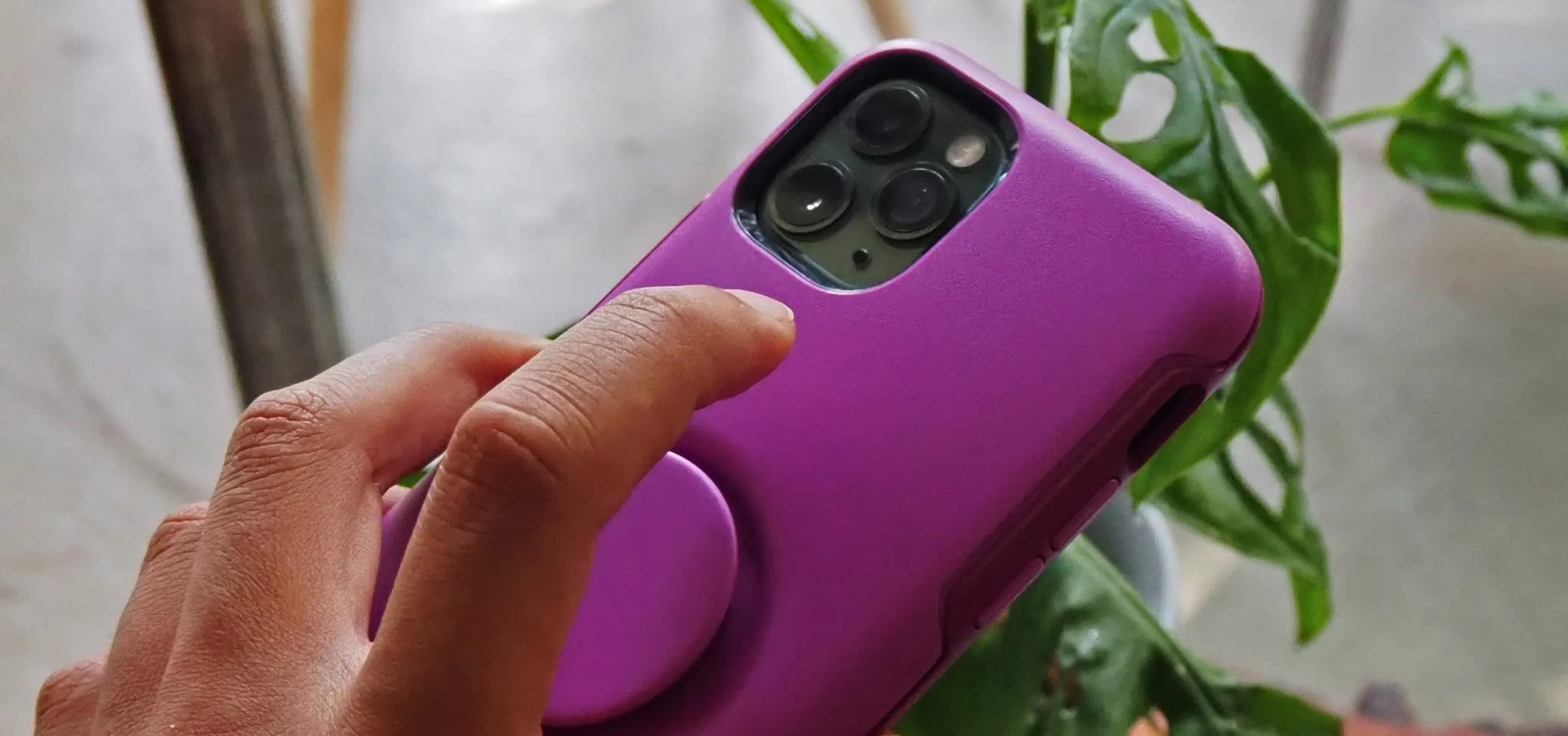Hand holding a purple phone case with a pop socket, positioned near a plant.