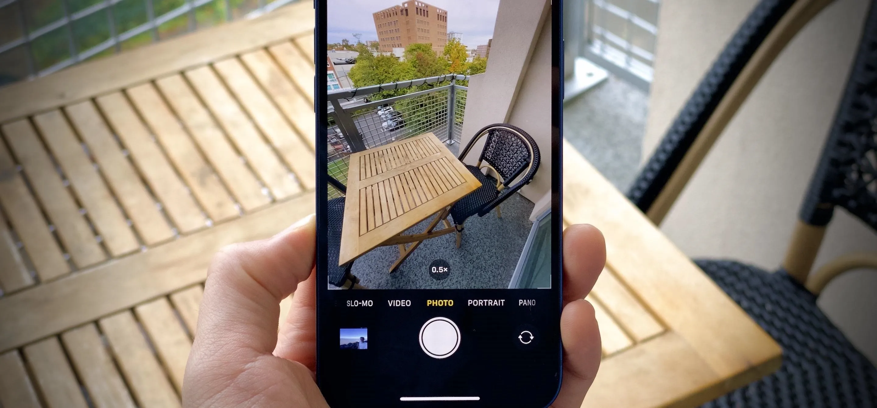 A hand holding a smartphone, capturing a photo of a table and a chair on a balcony.