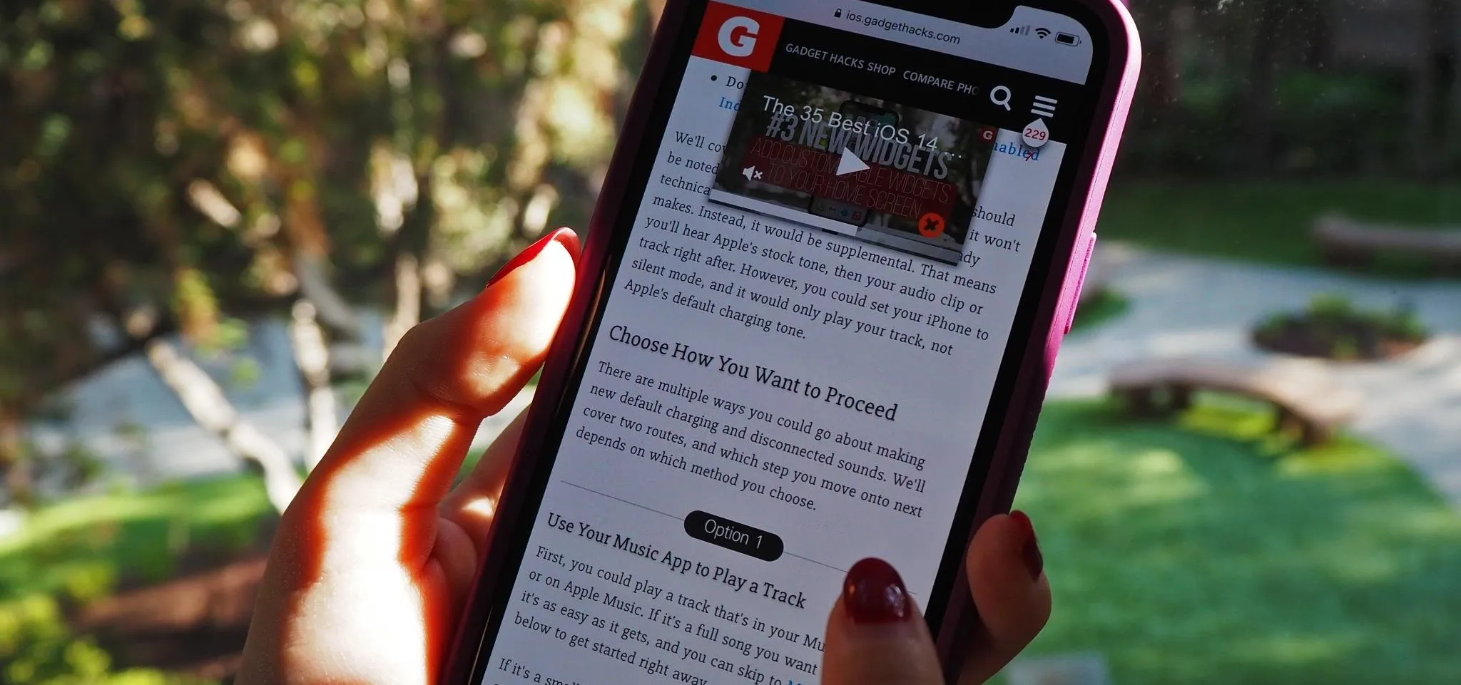 Person holding a smartphone displaying a web page with a video thumbnail, set against a blurred outdoor background.