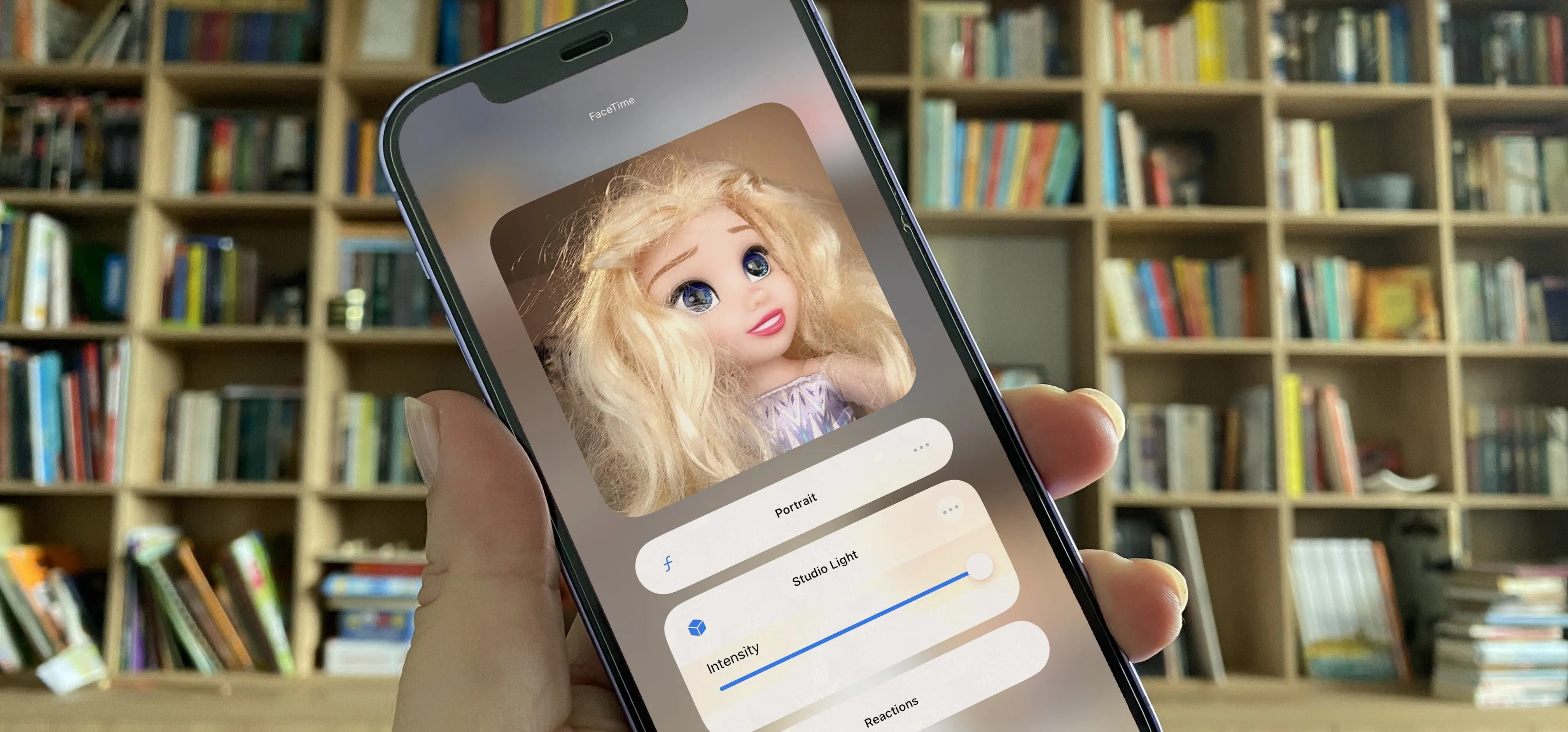 A phone displaying a doll with long blonde hair in front of a bookshelf.