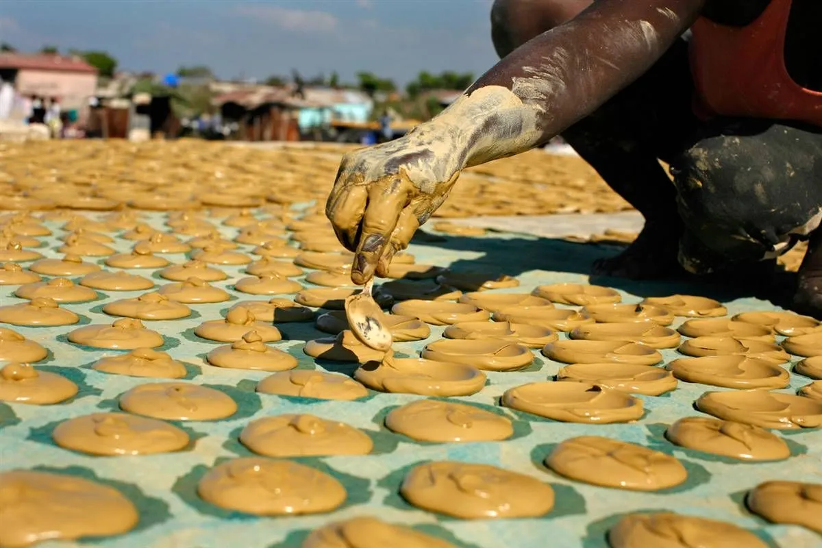 null A man pouring and shaping peanut-butter-looking clay into cookie shapes with hundreds of the dirt cookies, or mud cookies, are visible on the ground.