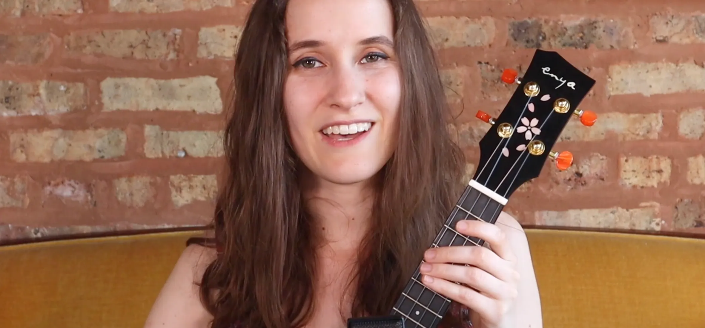 A woman with long hair holding a ukulele in front of a brick wall backdrop.