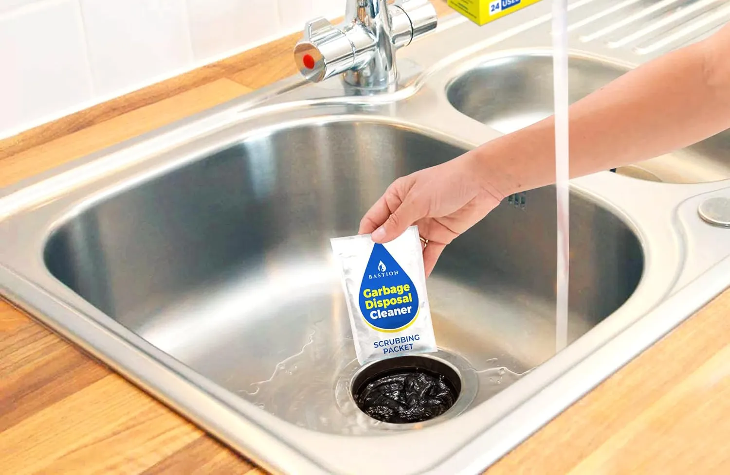 A person's arm placing a Bastion Garbage Disposal Cleaner biodegradable packet inside a garbage disposal in a kitchen sink with the water running.