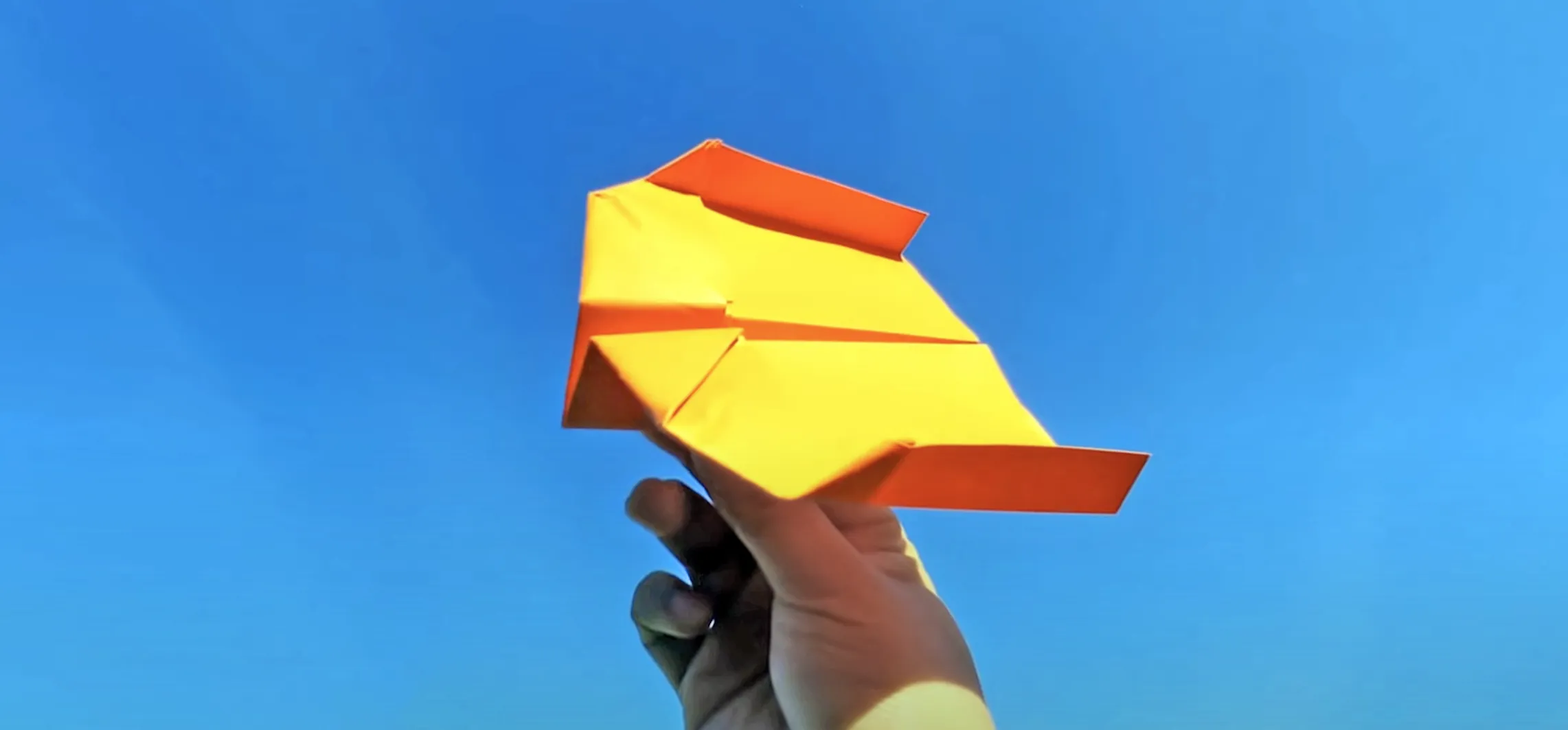 Hand holds orange boomerang paper airplane against cloudless blue sky, showcasing folded winglets designed for looping flight.