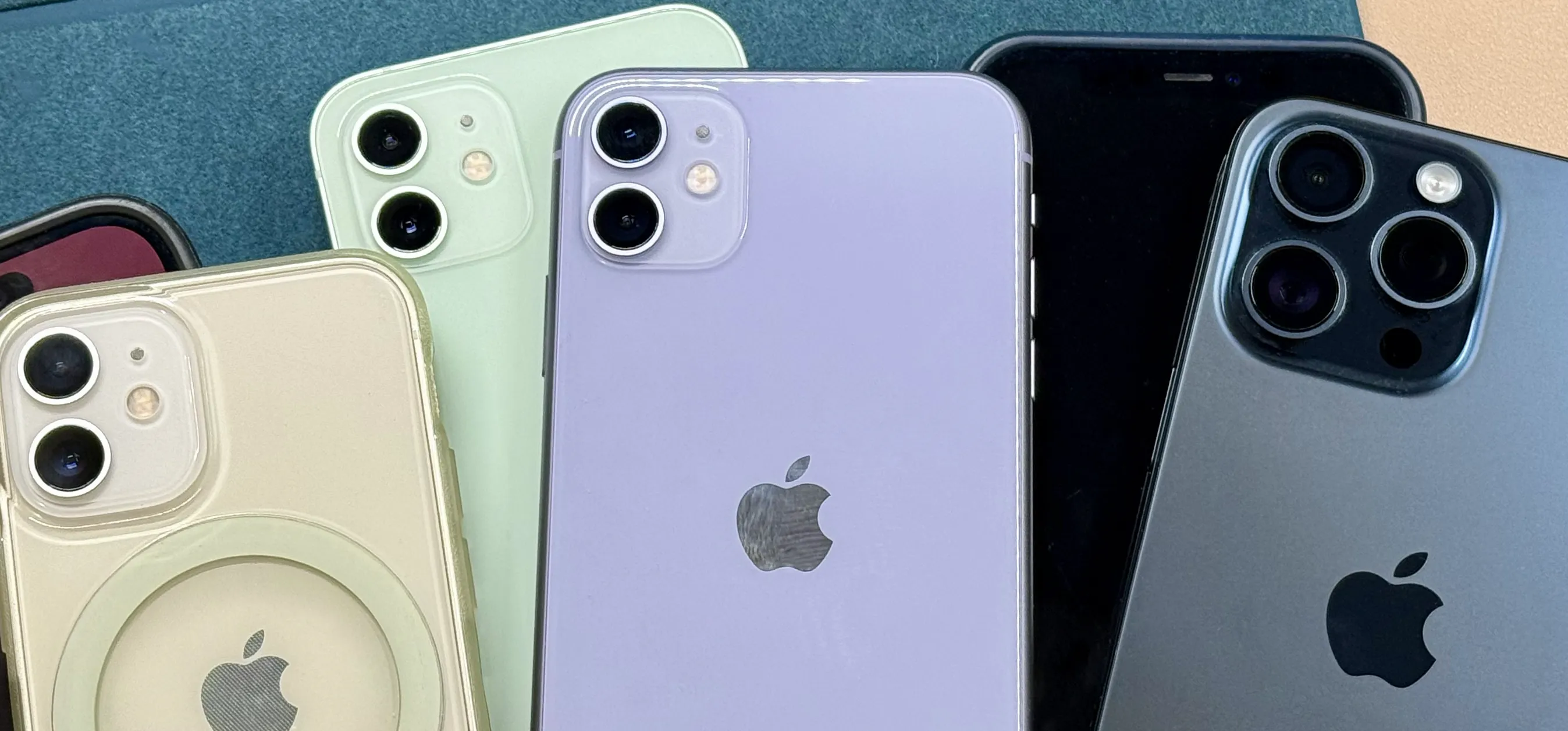 Six iPhones spread out on a green and tan surface. Two iPhones show the front displays, while the other four show the rears with cameras in different colors.