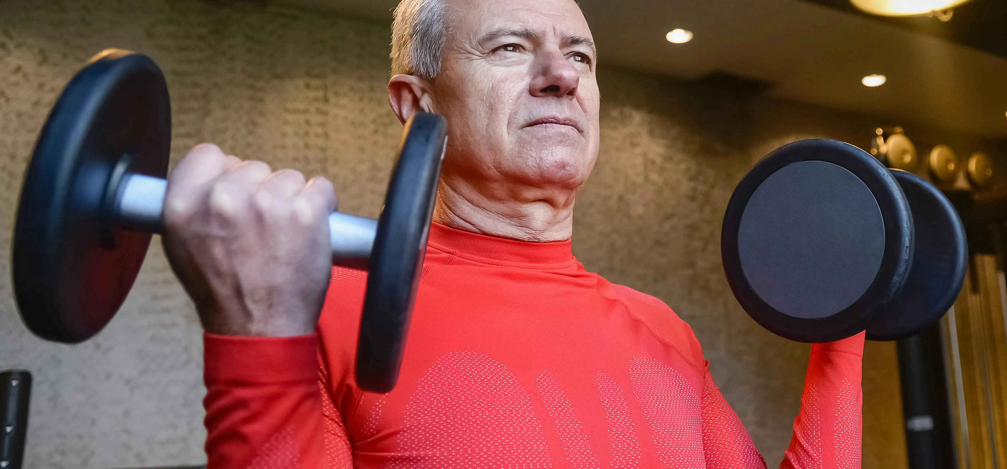 Senior man in red compression shirt performs dumbbell bicep curls indoors, focused expression, showcasing strength training and active aging fitness.
