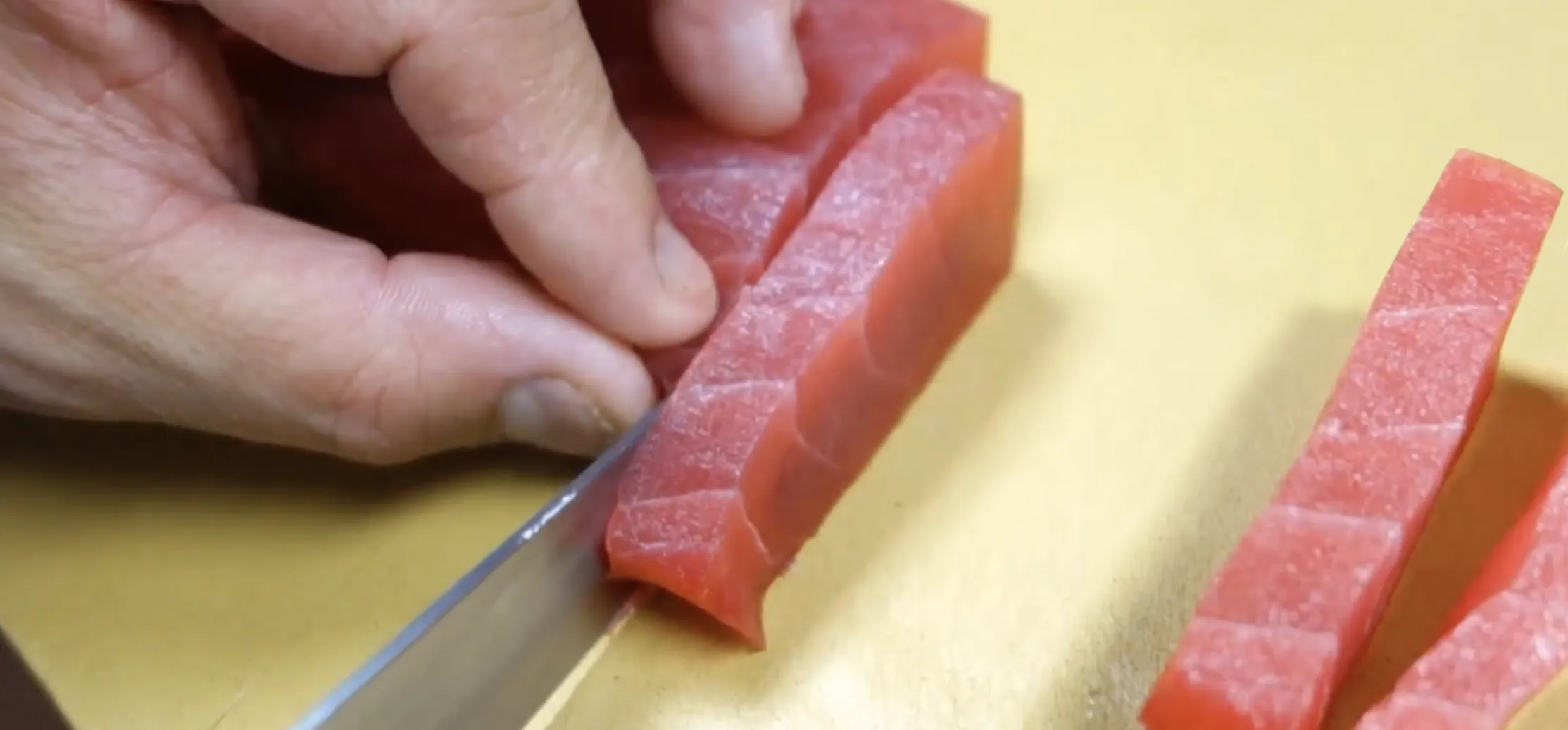 Hand holding bright red raw tuna on a cutting board, with a knife cutting medium-size slices for sashimi.