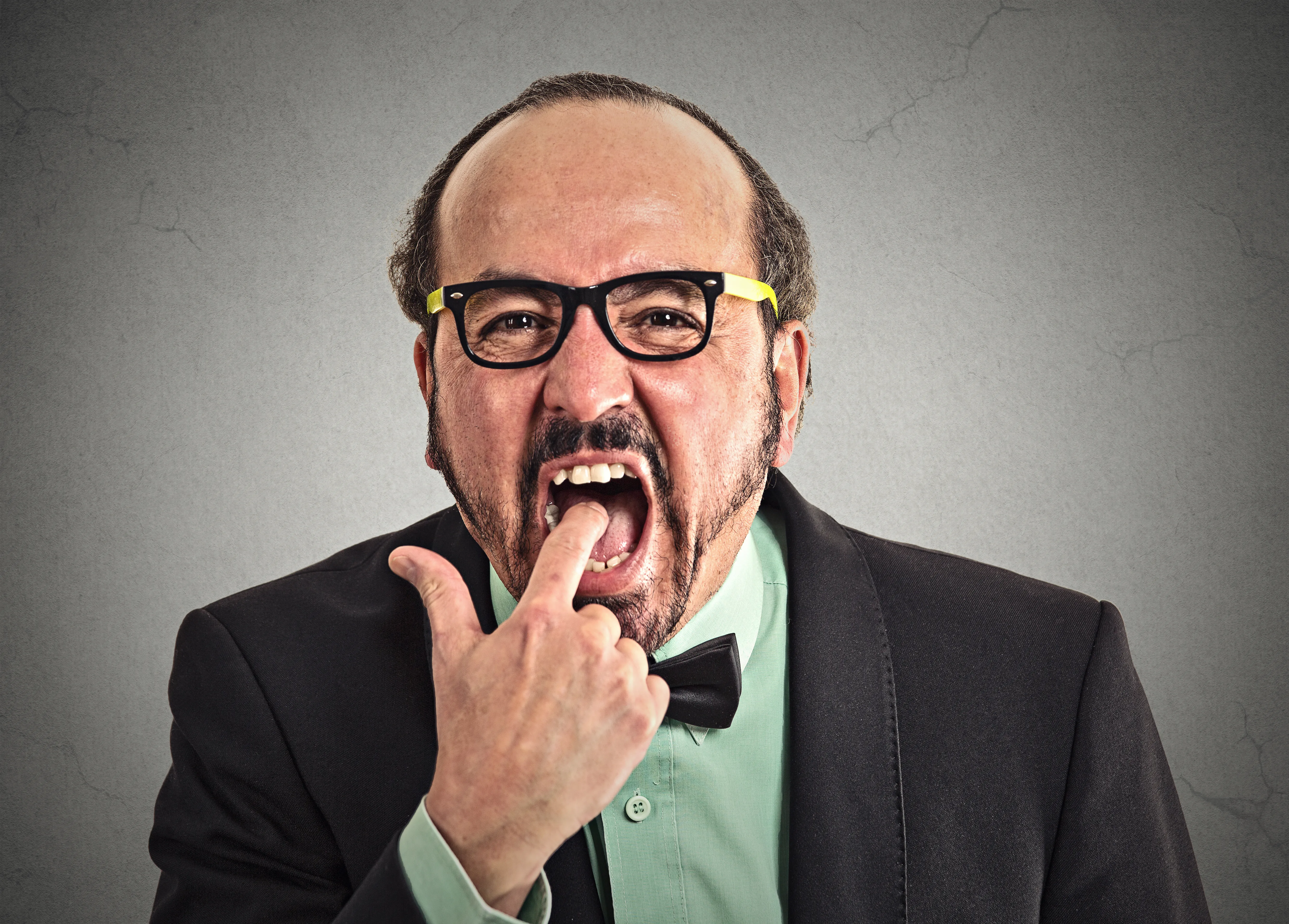 null A man in a business suit and green button-down shirt, wearing black glasses with yellow arms, points his finger into his mouth.