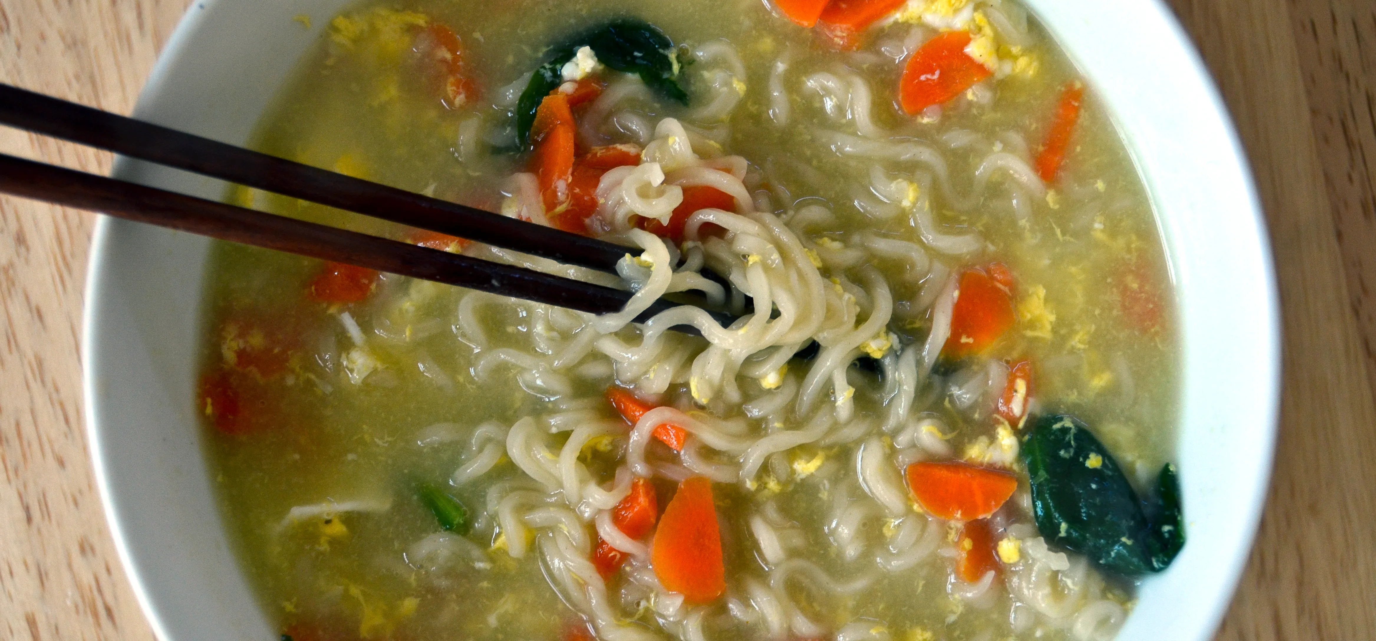 White bowl of ramen noodle soup with vegetables and chopsticks on a light-brown table.