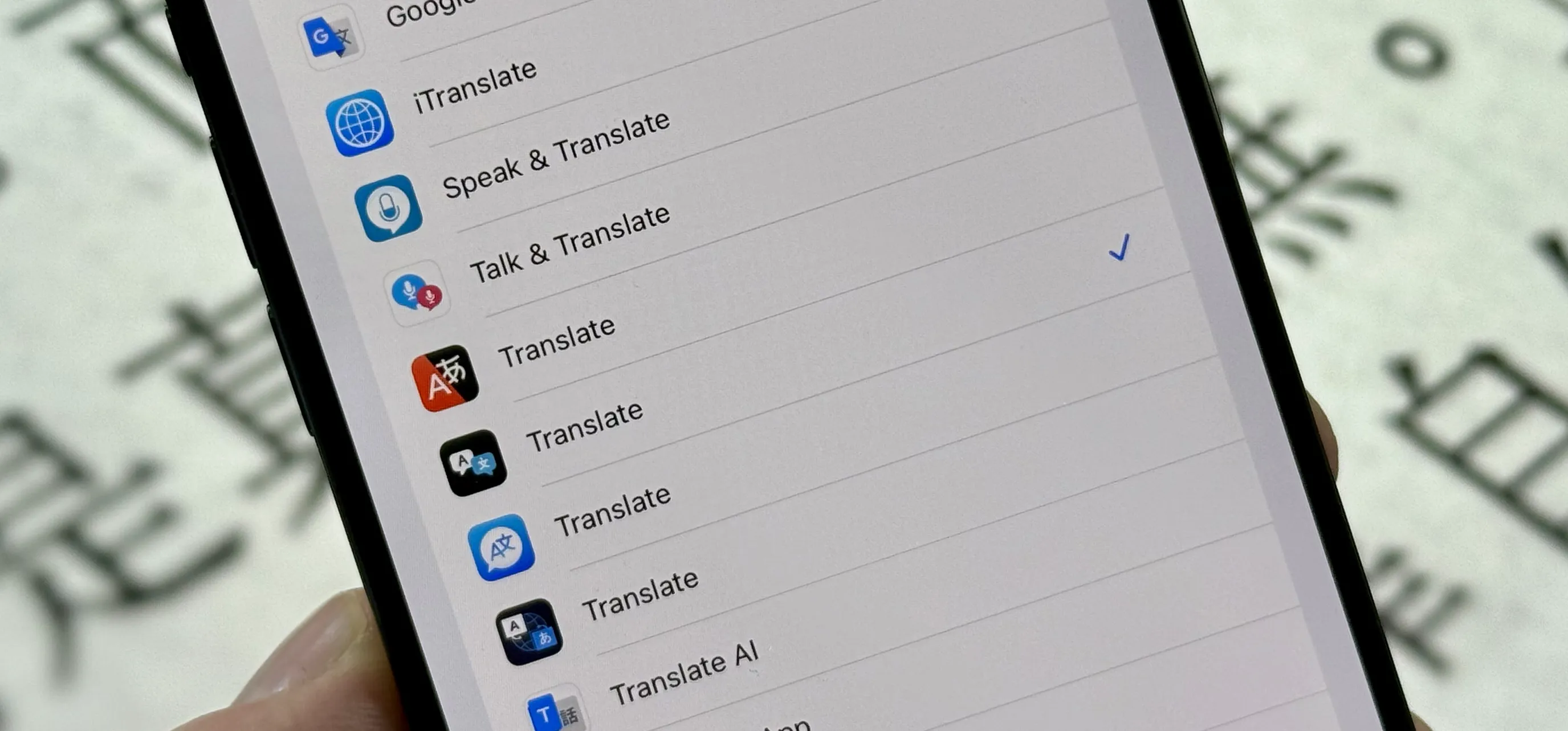 A smartphone screen displaying a list of translation apps, with “Talk & Translate” selected. The background is blurred, showing Japanese characters on a printed surface.