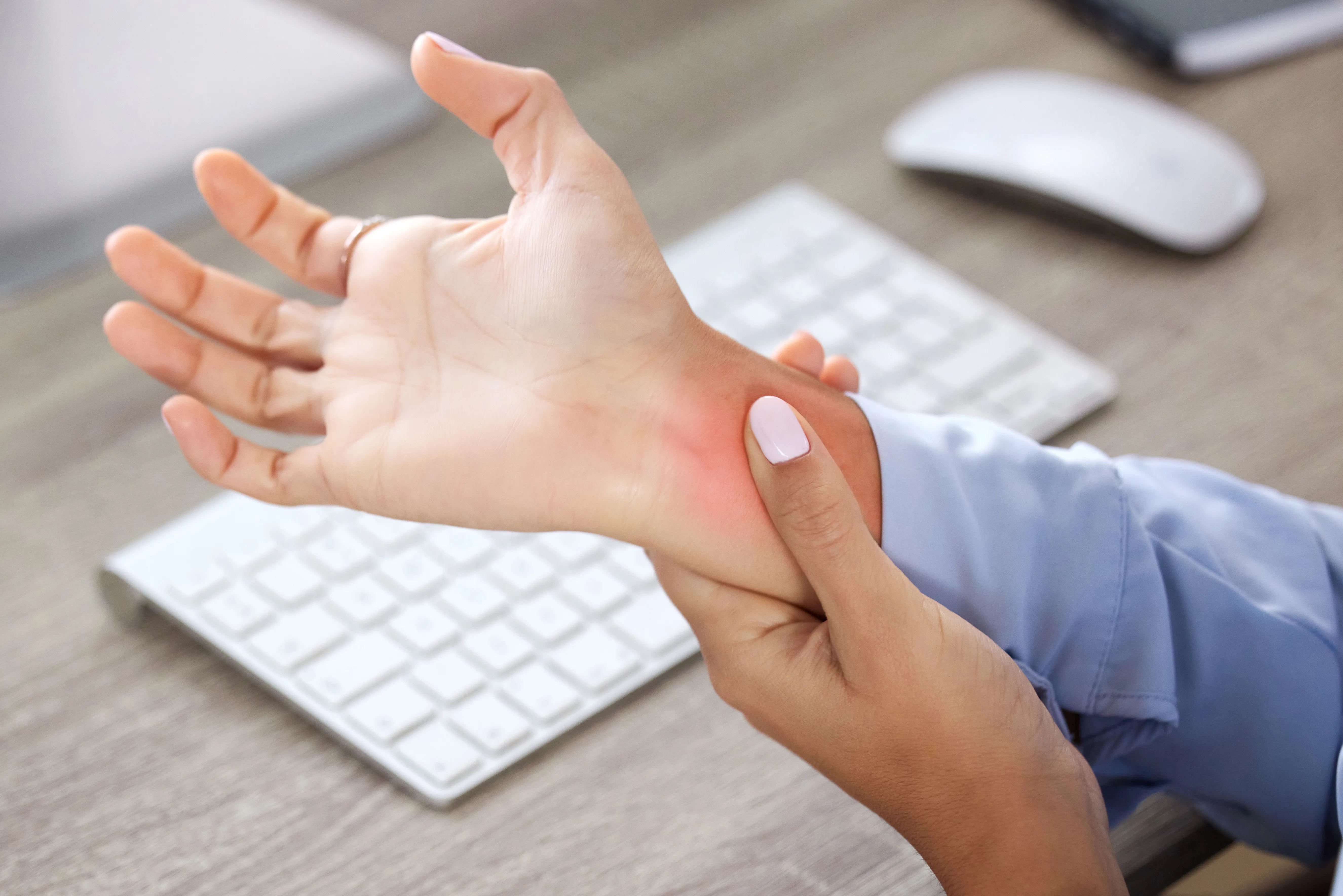 Unrecognizable business woman sitting alone at her desk, holding her forearm above her wrist using her left hand; a computer keyboard and mouse are on the desk.