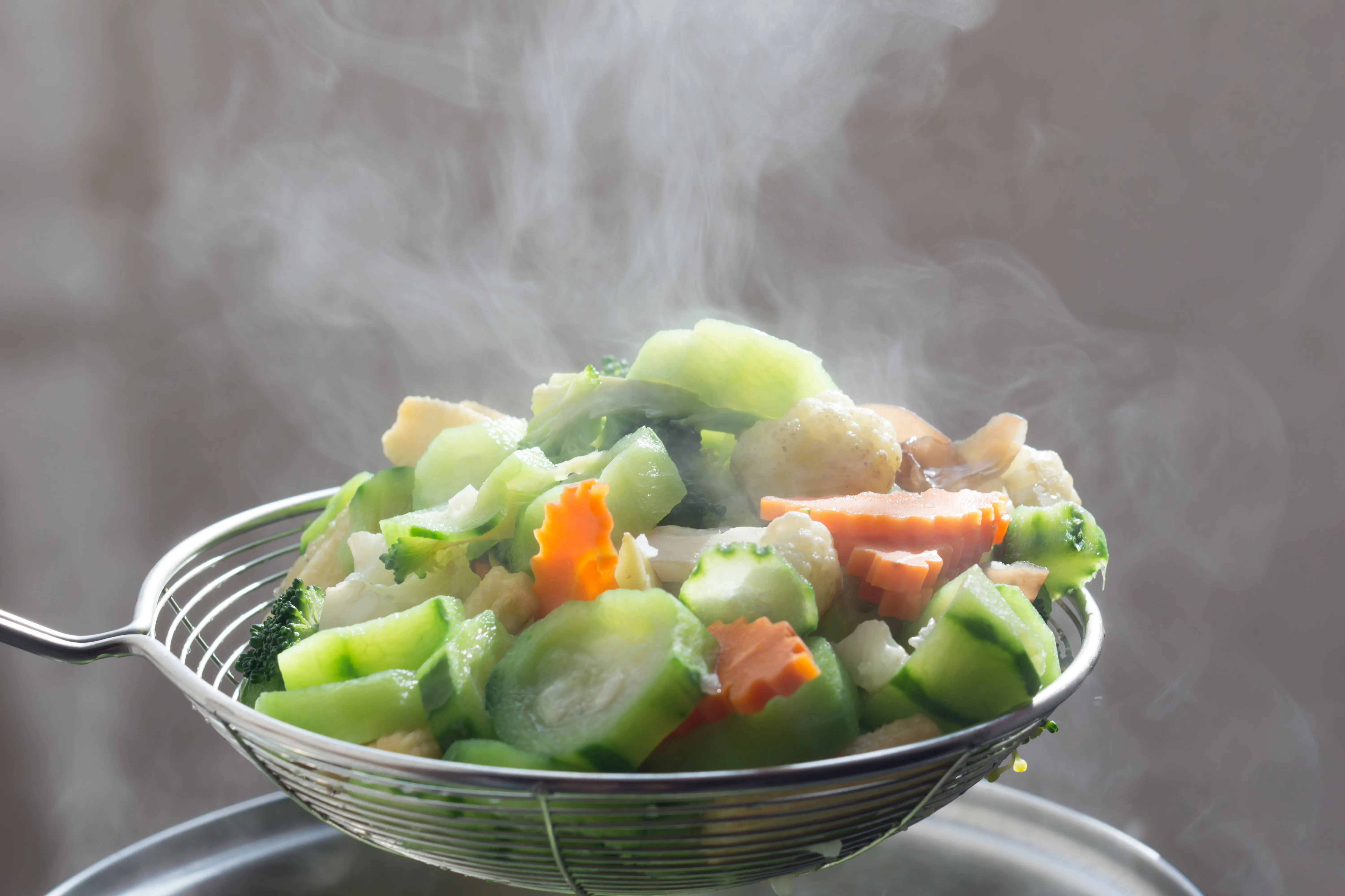 A wire colander holding steaming hot cut veggies above a pot.
