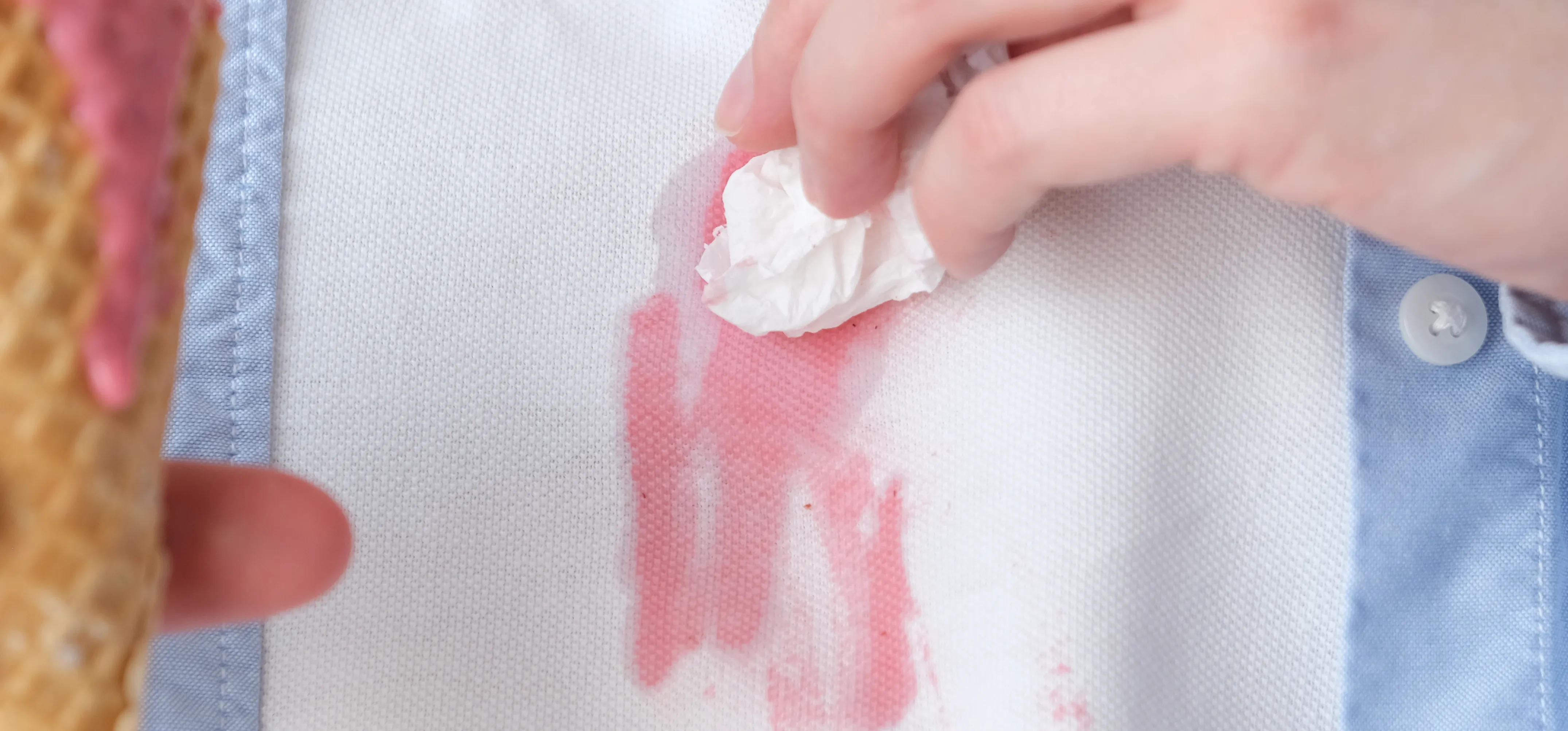 Man blotting a pink ice cream stain on his white shirt; he's holding a waffle cone and dripping pink ice cream in the foreground with his other hand.