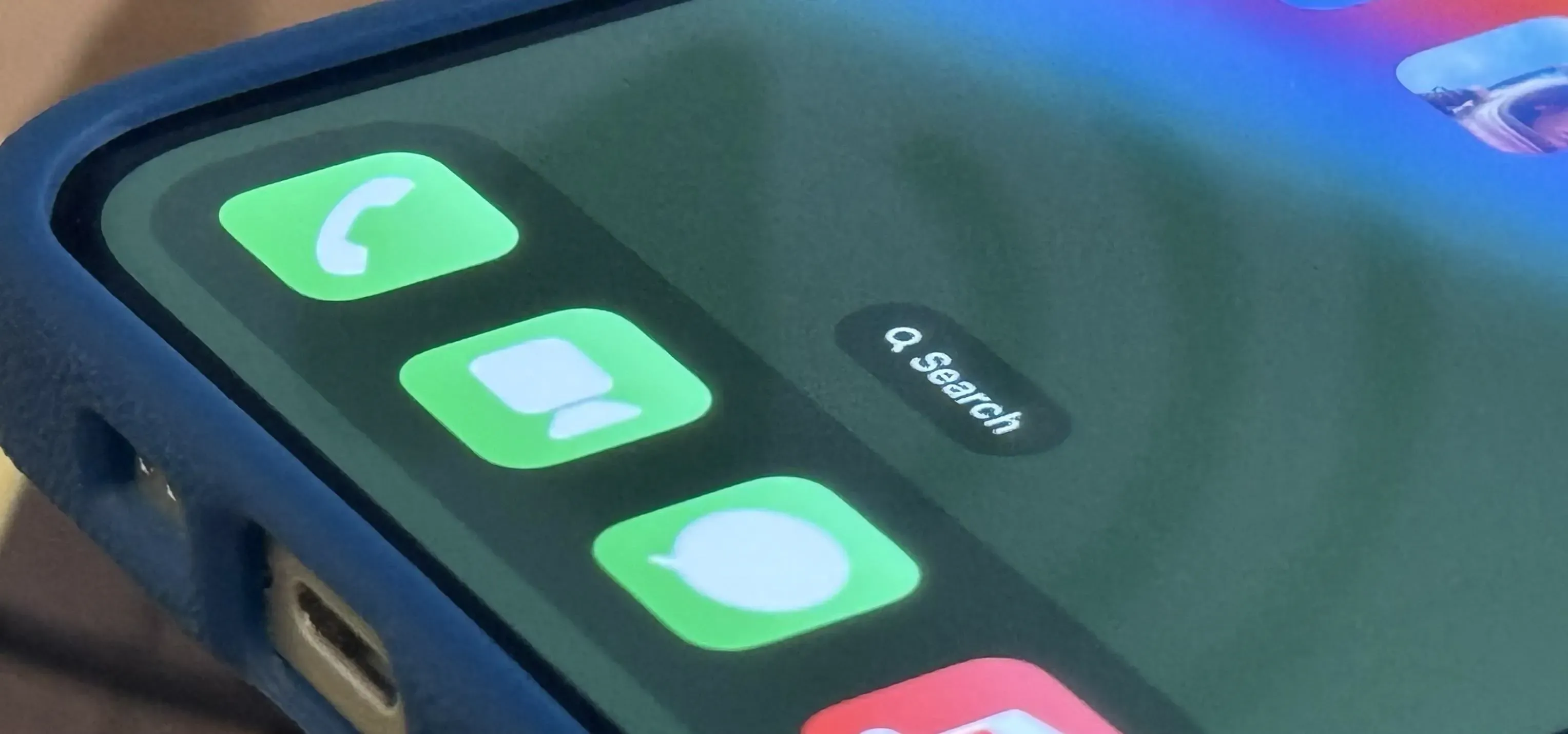 Close-up of iPhone home screen corner showing green Phone, FaceTime, Messages icons and a Search widget bubble along the screen edge; a speaker playing icon can be seen in the reflection.