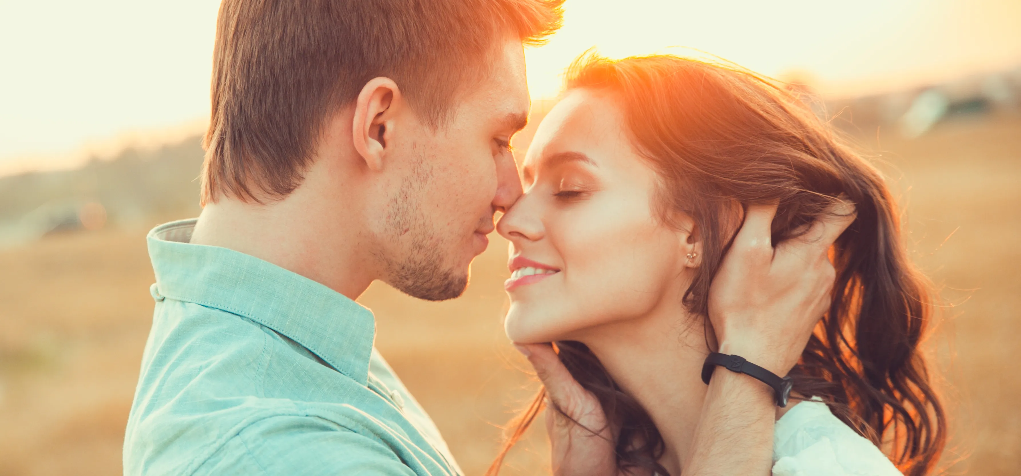 Couple stands close at sunset, eyes closed, smiling softly, about to kiss while gently holding each other's heads.