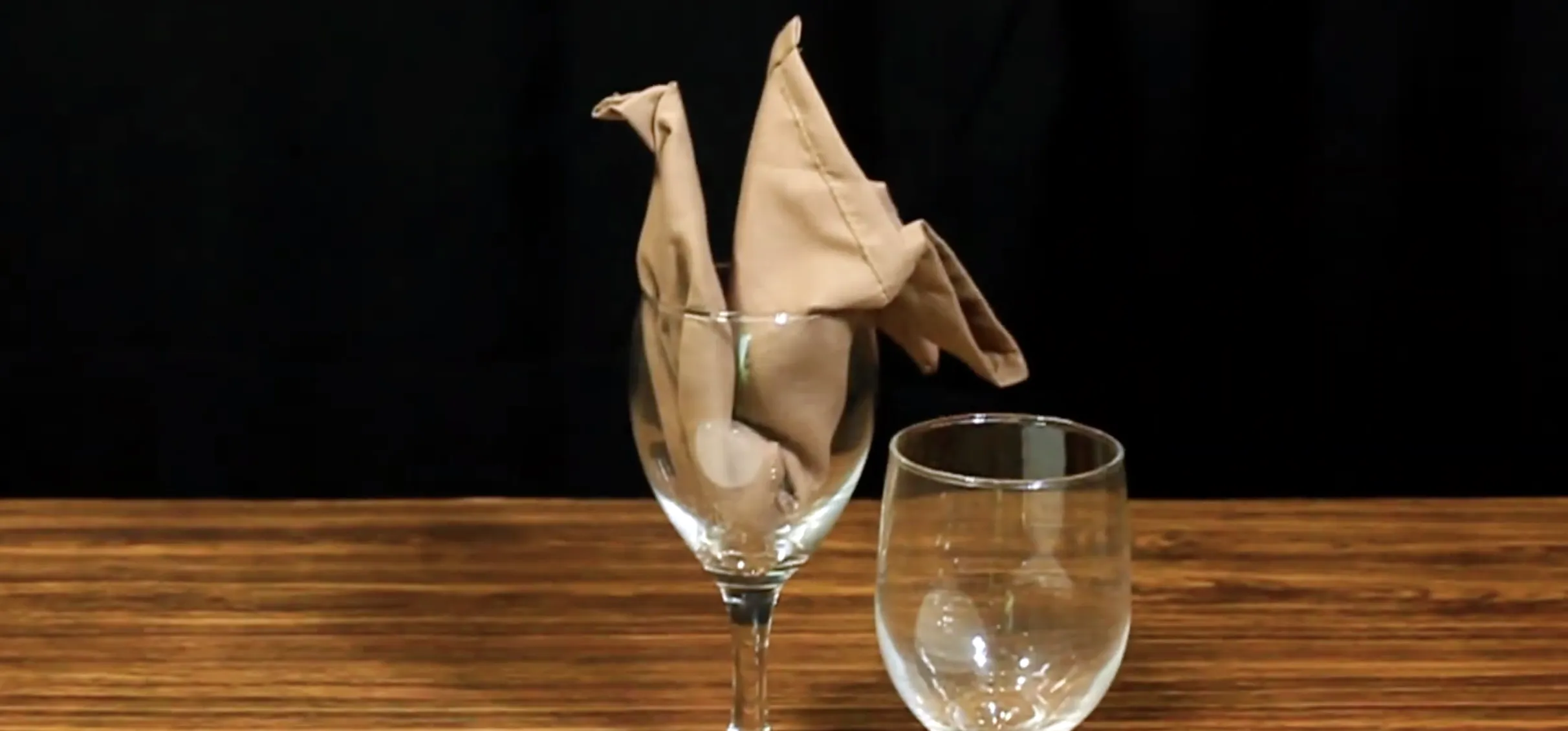 Napkin folded swan or peacock-like bird displayed in a wine glass on a table setting.