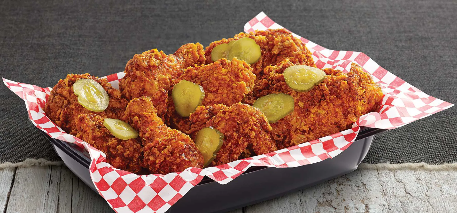 null Eight-piece Nashville hot sauce fried chicken meal in a to-go container with white-red checkered paper lining and pickles on top.