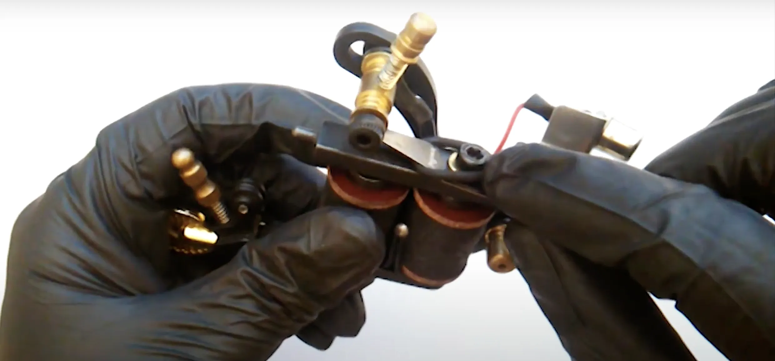 Black-gloved hands fit rubber O-ring onto coil tattoo machine armature bar, stabilizing contact screw.