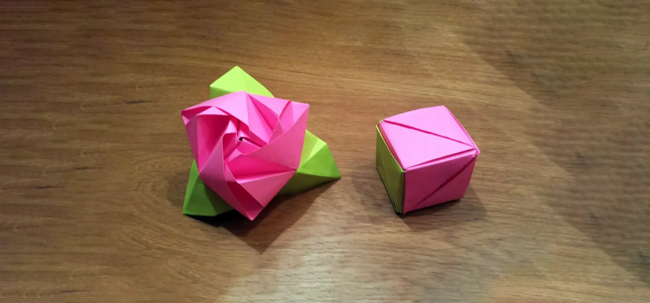 Pink modular origami rose with green leaves sits beside matching cube model on polished wood surface.