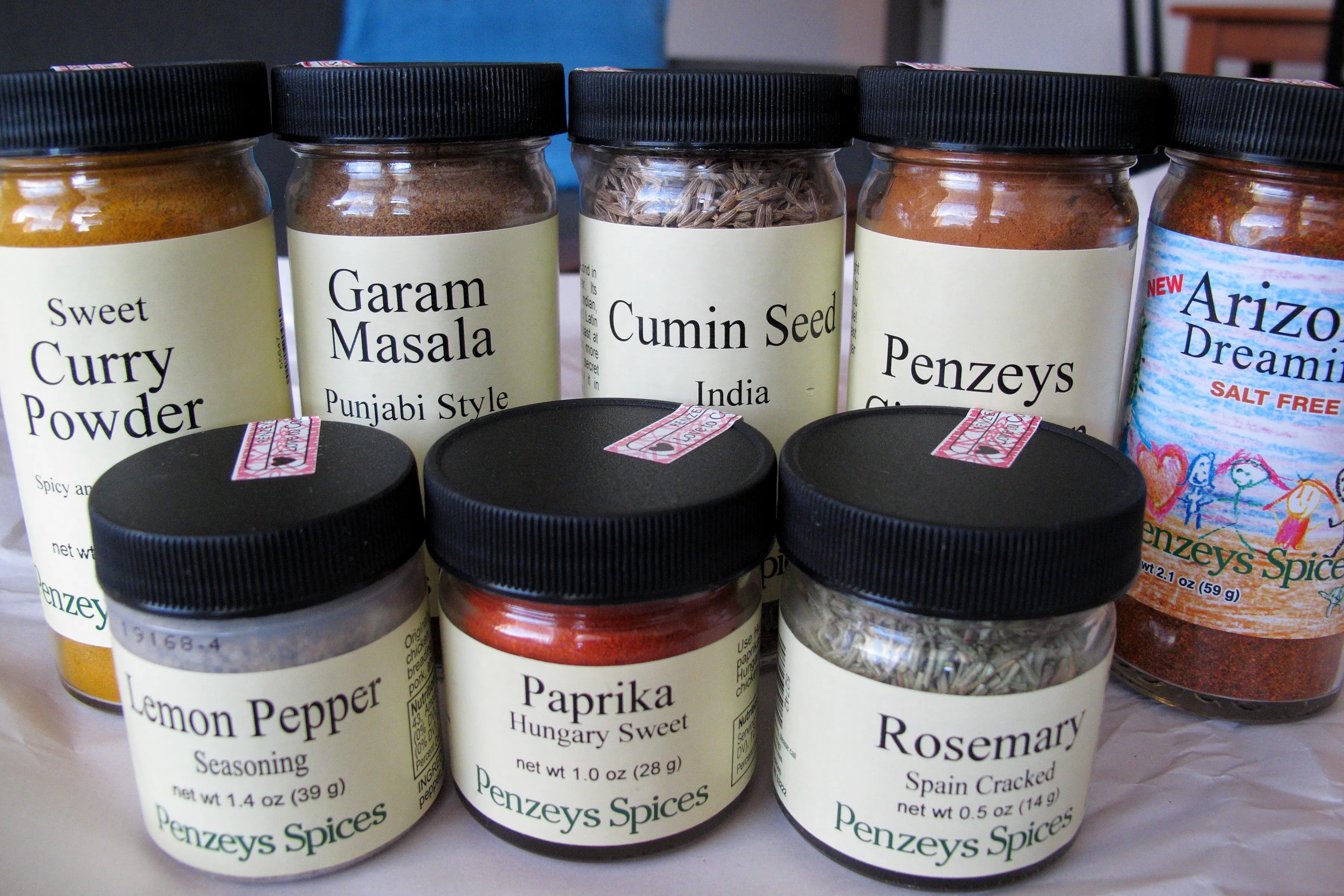 null Row of spice jars (sweet curry powder, garam masala, cumin seeds, Penzeys, Arizona Dreaming) and another a row of smaller jars below it (lemon pepper, paprika, rosemary).