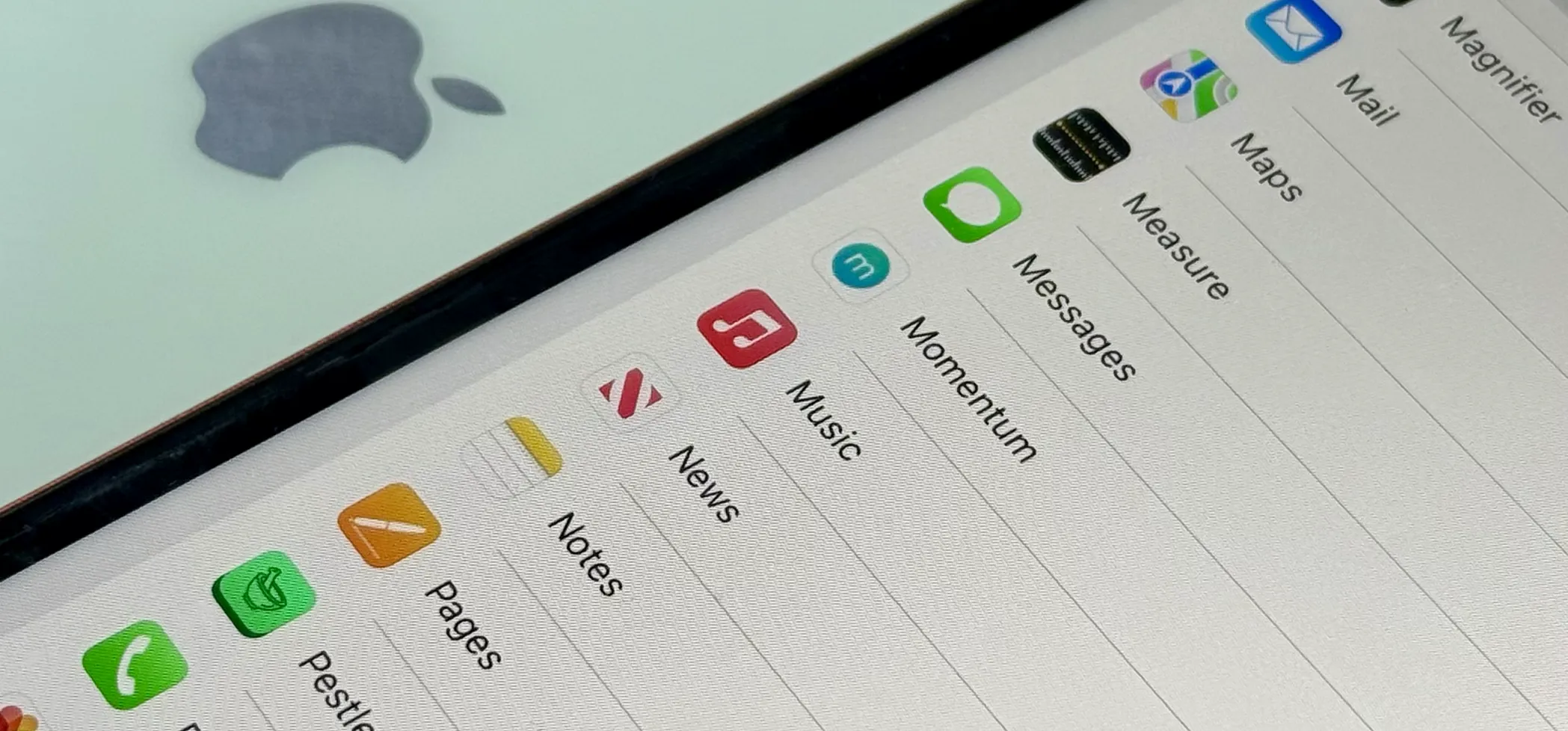 Close-up of iPhone screen showing app list in Settings with Messages, Music, News, and other icons, positioned over a silver Apple logo on another iPhone.