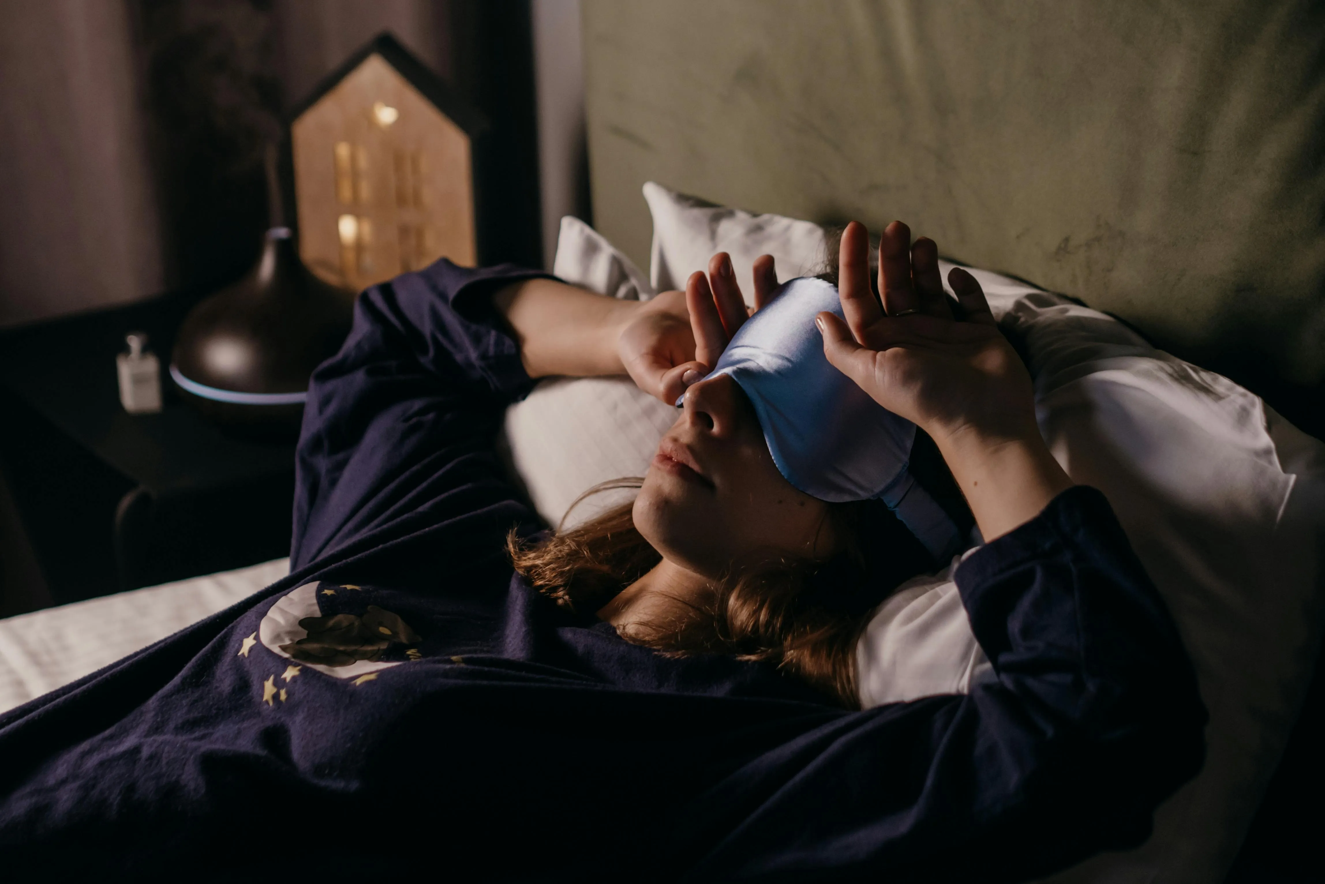 Woman in navy pajamas adjusts silky blue sleep mask while reclining in bed — aroma diffuser and nightlight glowing nearby.