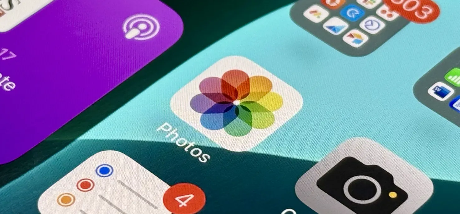 Close-up of iPhone Home Screen highlighting Apple’s multicolored Photos app icon amid other app icons, including Camera, Reminders, Podcasts, and a folder with unread badges.