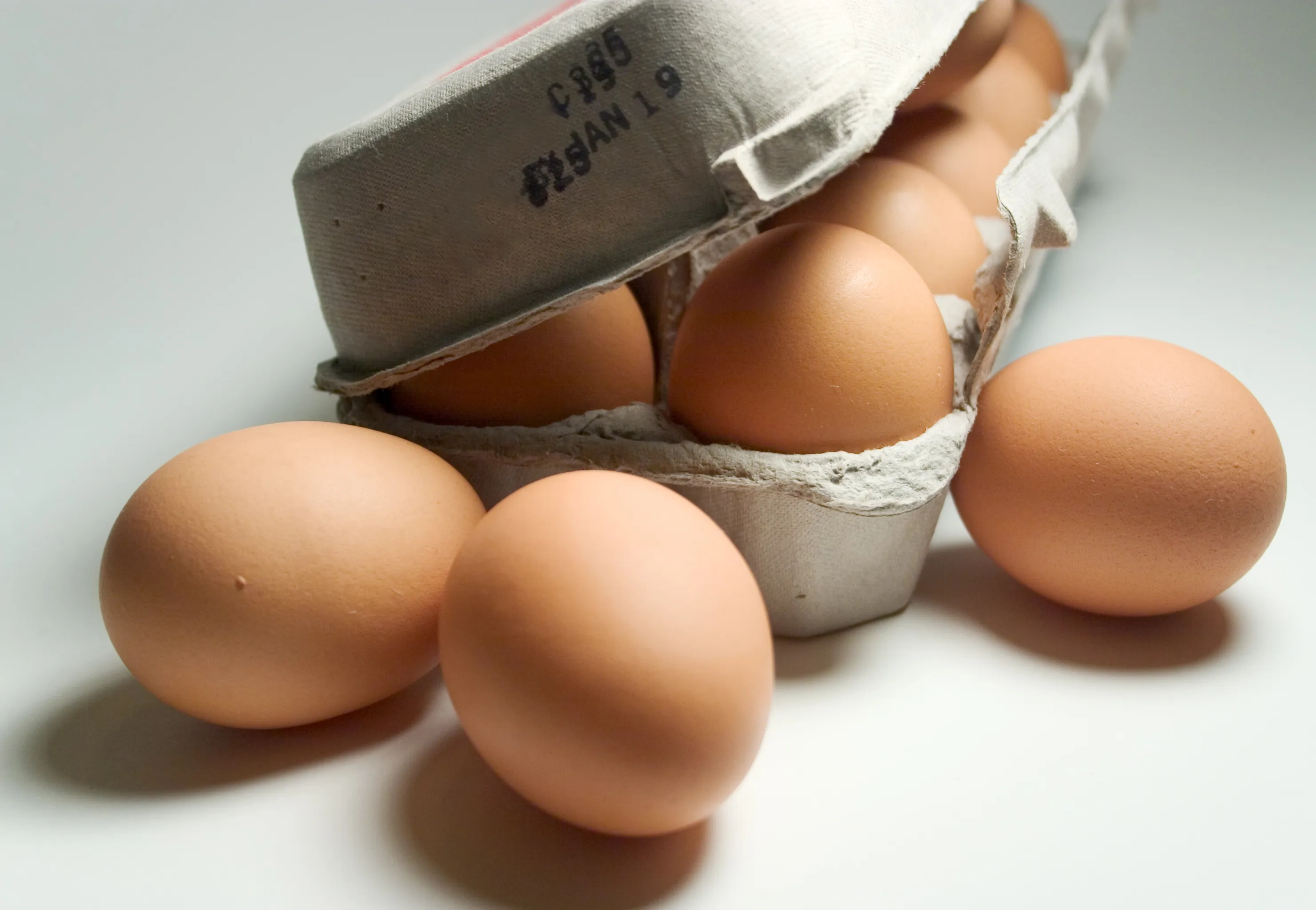 A carton of brown raw eggs with the lid open slightly and three eggs on the table outside of the carton. On the carton lid, there is a stamped date.