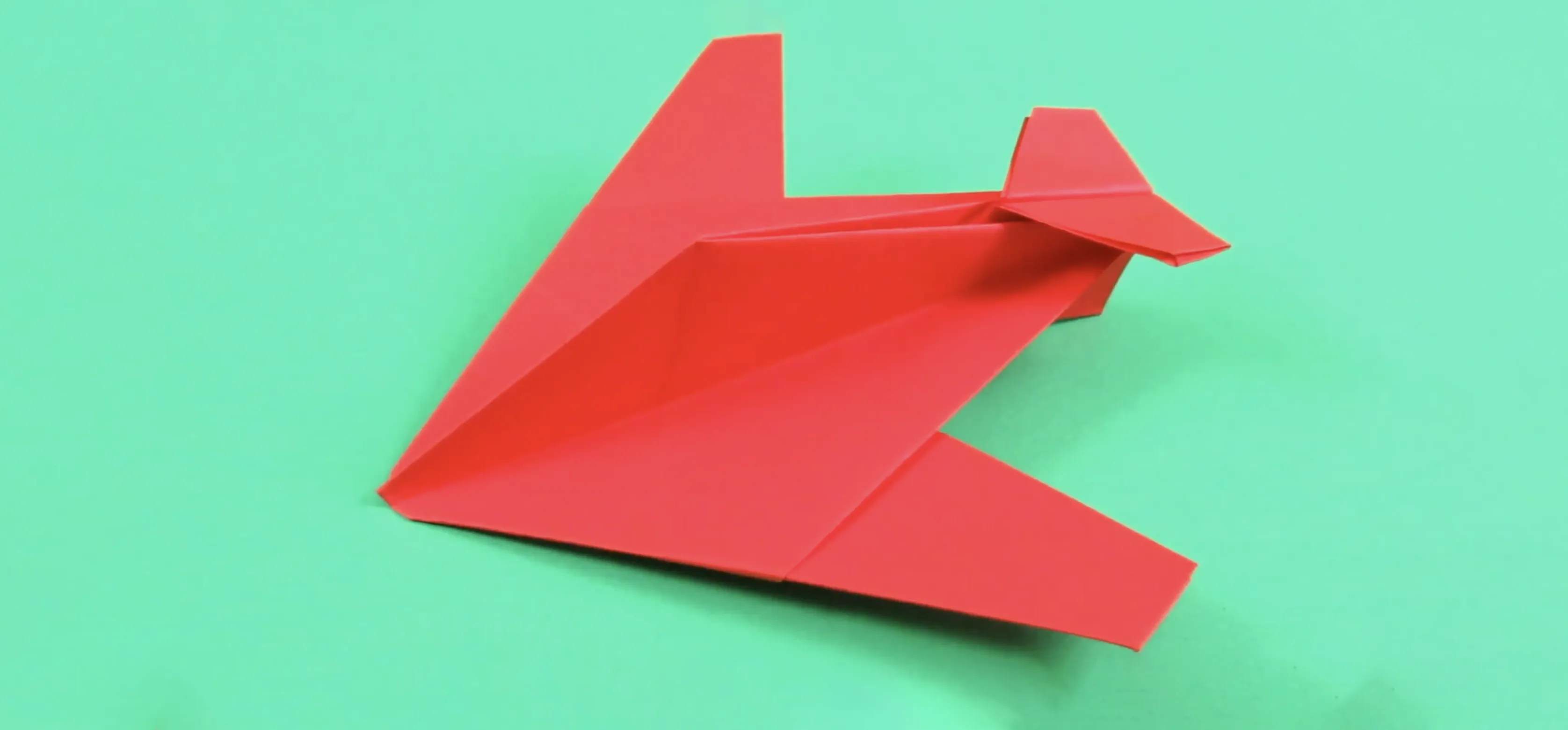 Red origami stealth fighter paper airplane rests on mint green surface, angular wings folded for aerodynamic look.