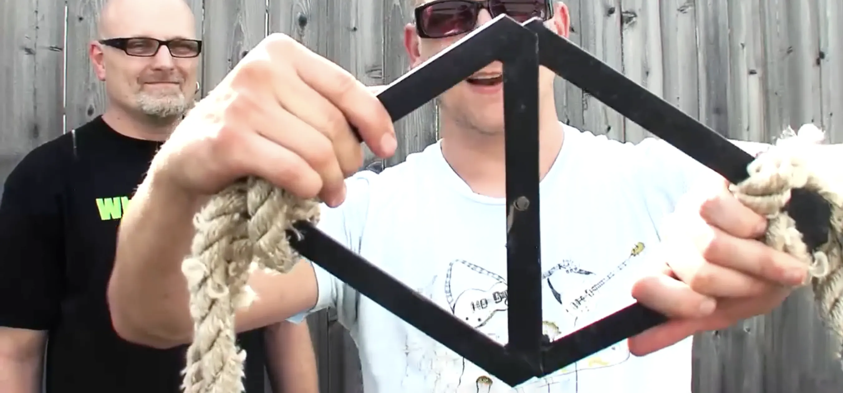 Man grips black metal diamond handle tied to rough rope for strongman training, friend wearing glasses watches near wooden fence.