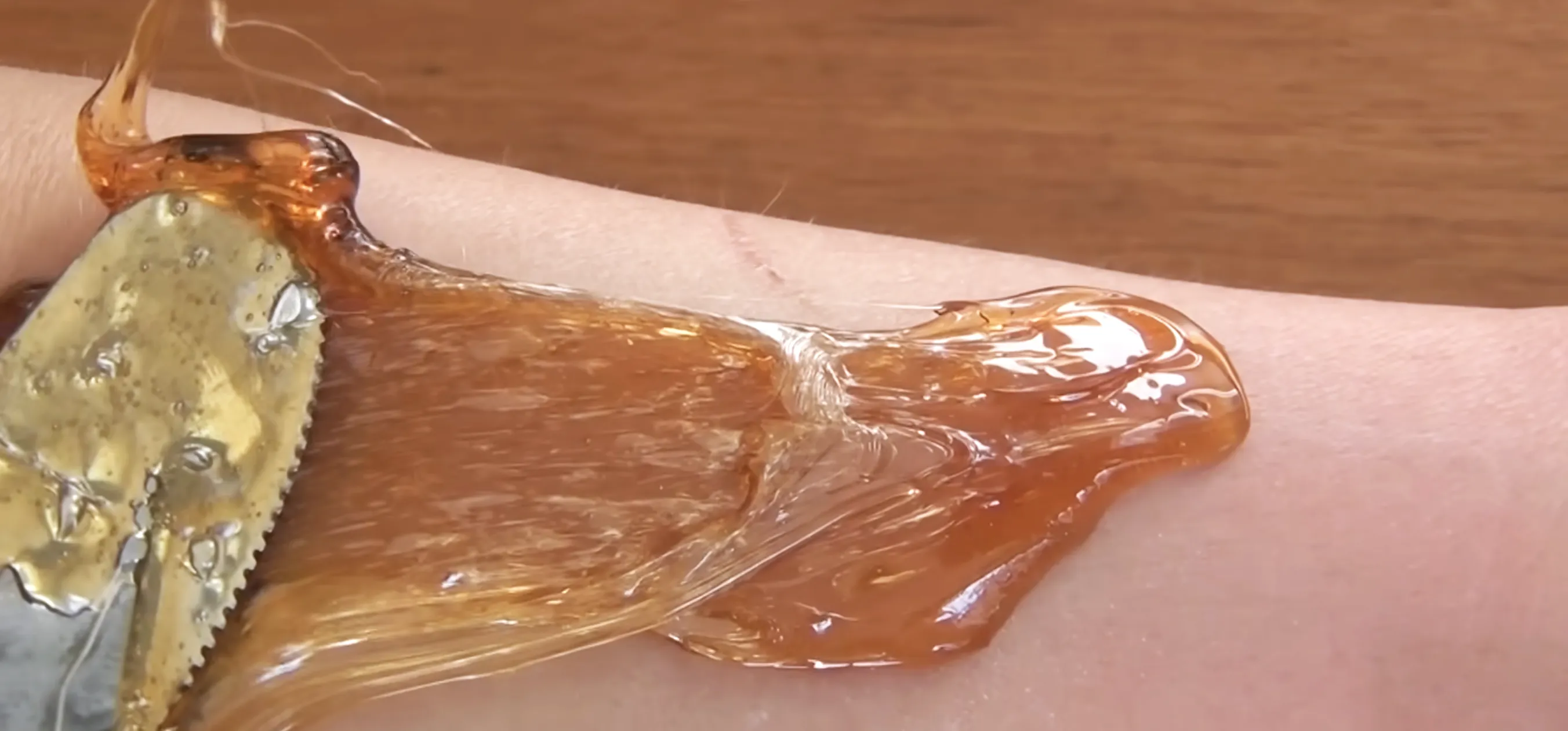 Amber sugar wax pulled with metal spatula across forearm, tugging fine hairs during at-home hair removal.