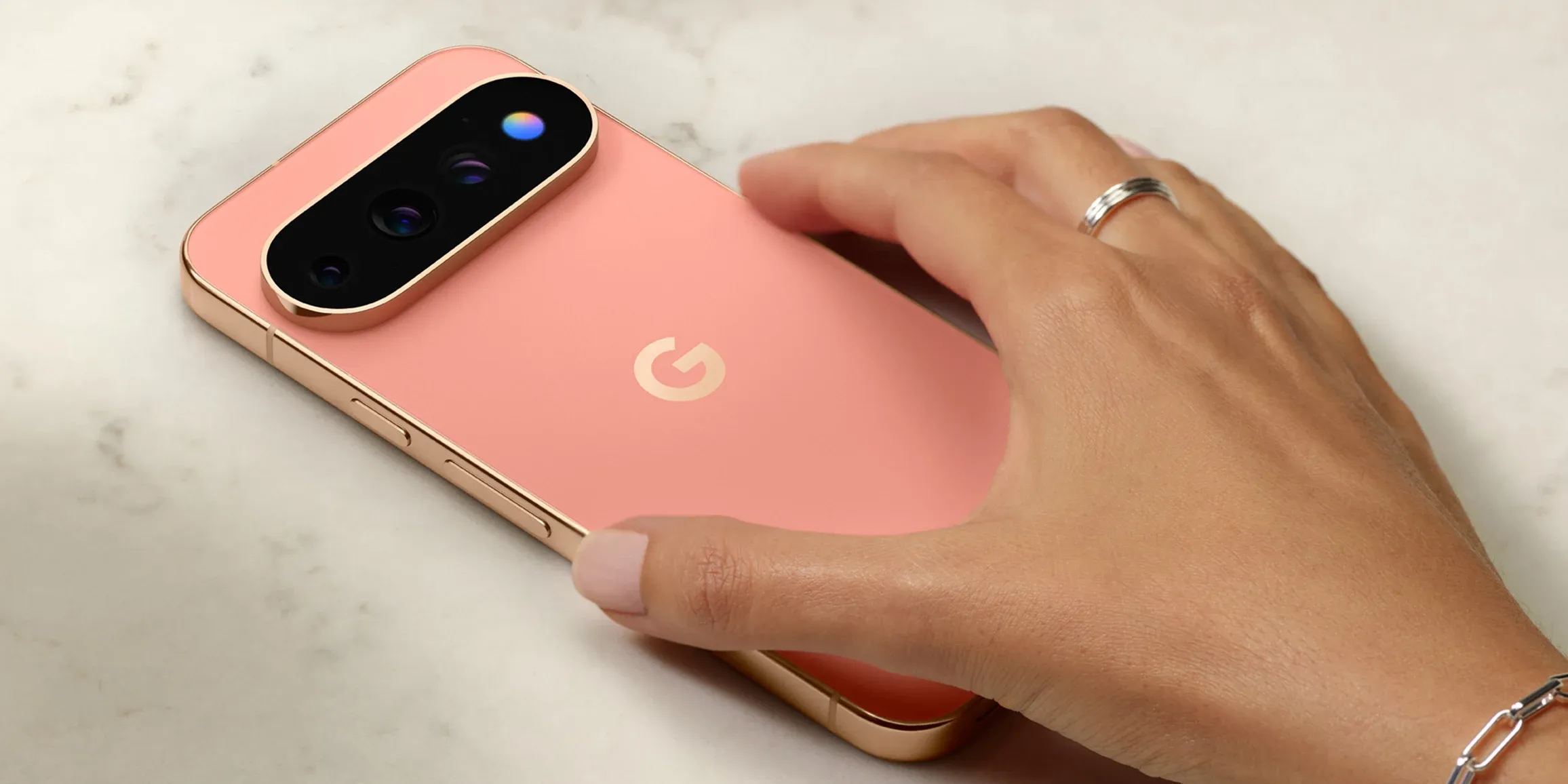 Google Pixel 11 in a coral finish resting on a marble surface beside a hand