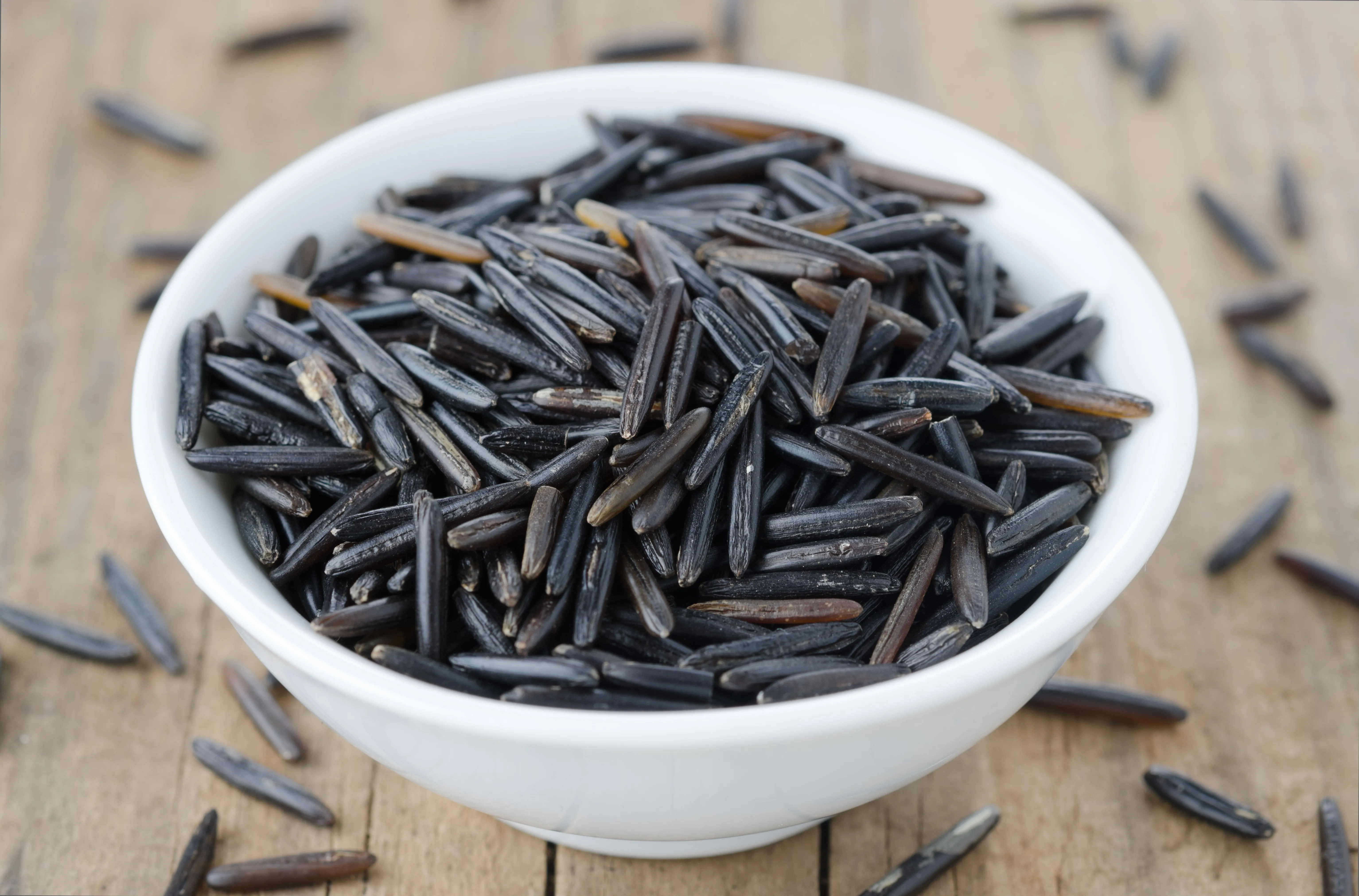 null Dark, black wild rice in a white ceramic bowl with grains on the table below.