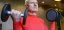 Senior man in red compression shirt performs dumbbell bicep curls indoors, focused expression, showcasing strength training and active aging fitness.