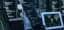Female hacker holds a smartphone displaying source code in front of multiple monitors showing terminal windows and a world map in a dark cybersecurity workspace.