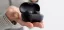 A person holds an open matte black wireless earbuds case in their palm, revealing the earbuds inside against a blurred background and striped long-sleeve shirt.
